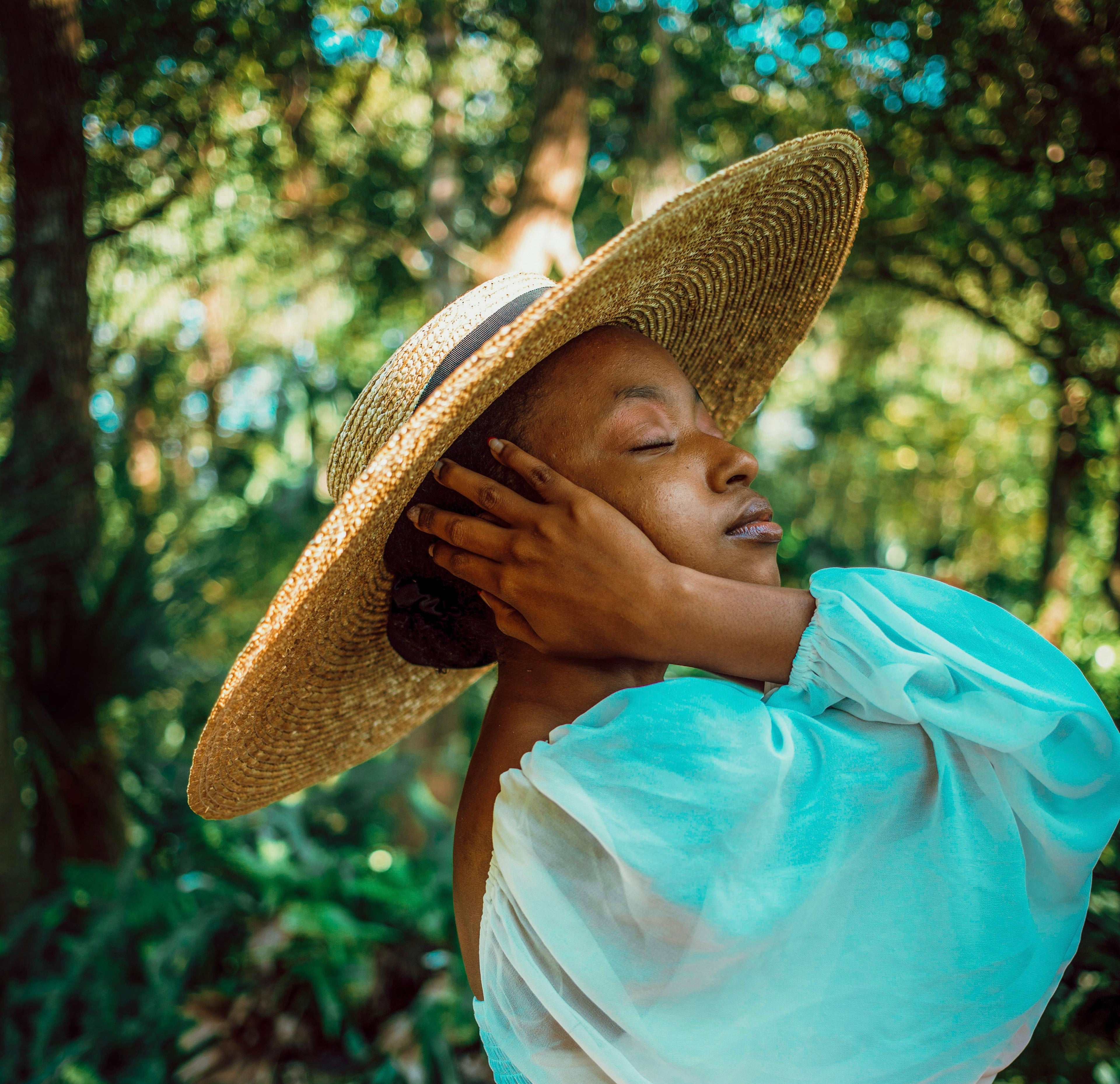A woman outdoors in a sunlit forest setting, wearing a large straw hat, a sheer light aqua puff-sleeve top, and an orange patterned bottom. Her eyes are closed and her hand rests against the brim of the hat.